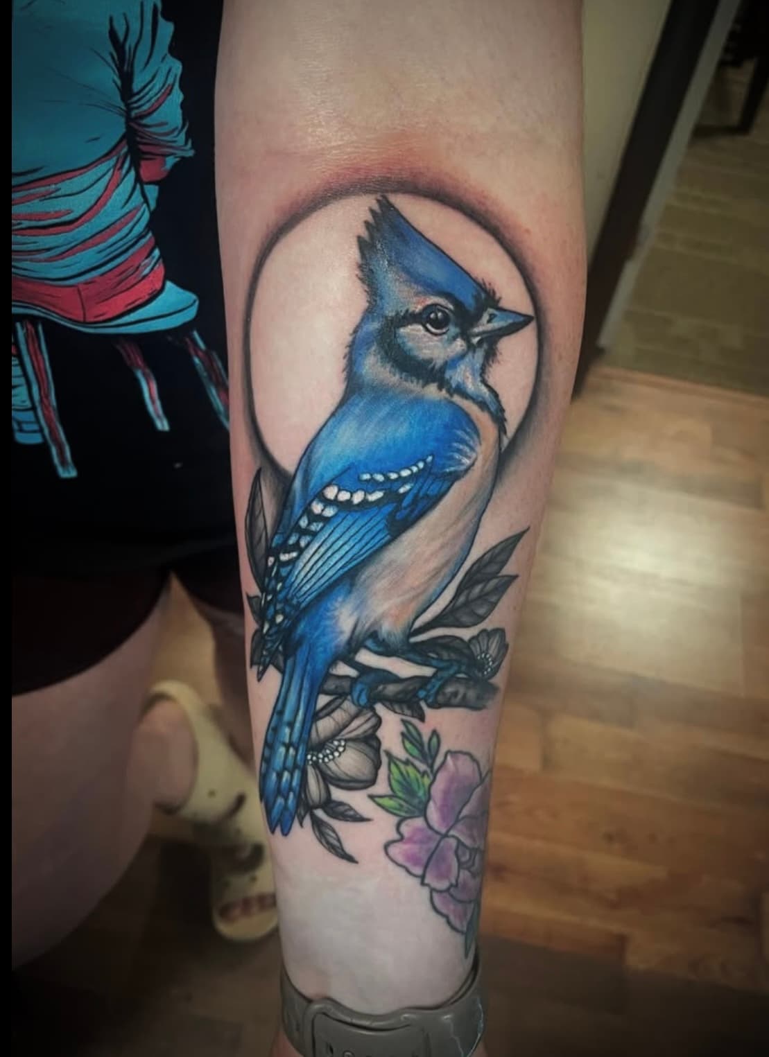 Blue jay tattoo with flowers on leg, perched on branch with circular background