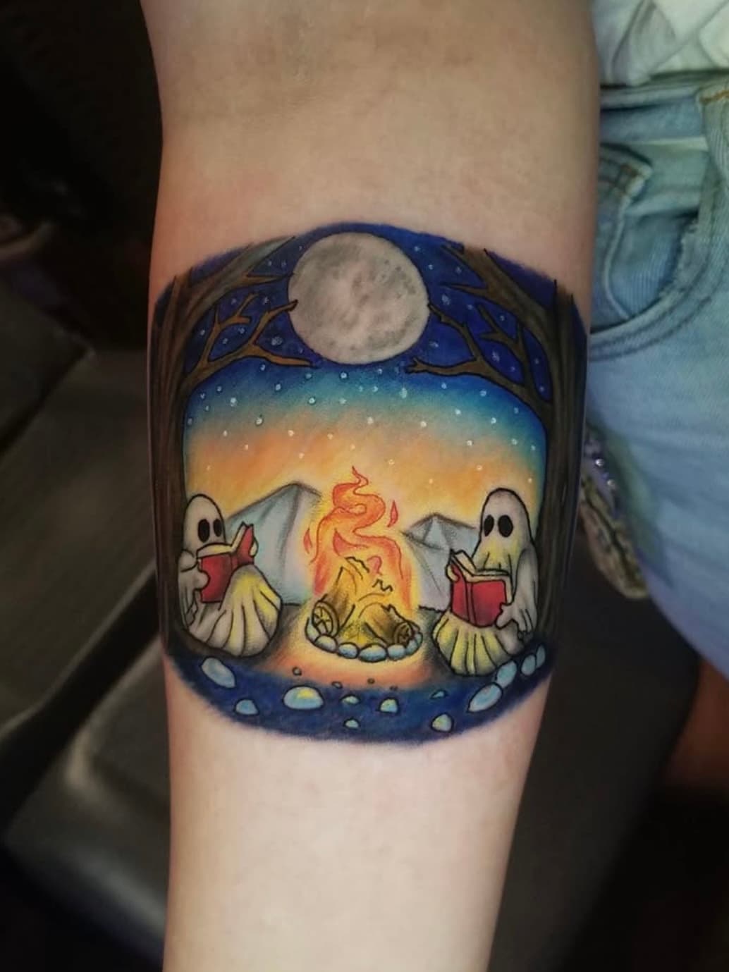 Colorful tattoo of two ghosts reading by a campfire under a full moon