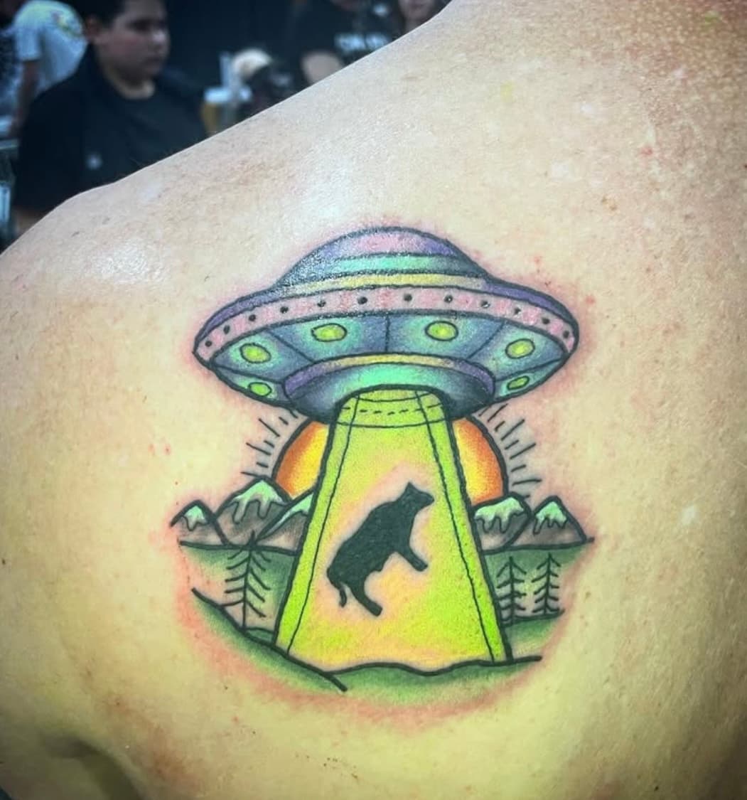 Colorful UFO tattoo with cat silhouette in beam of light above pine trees