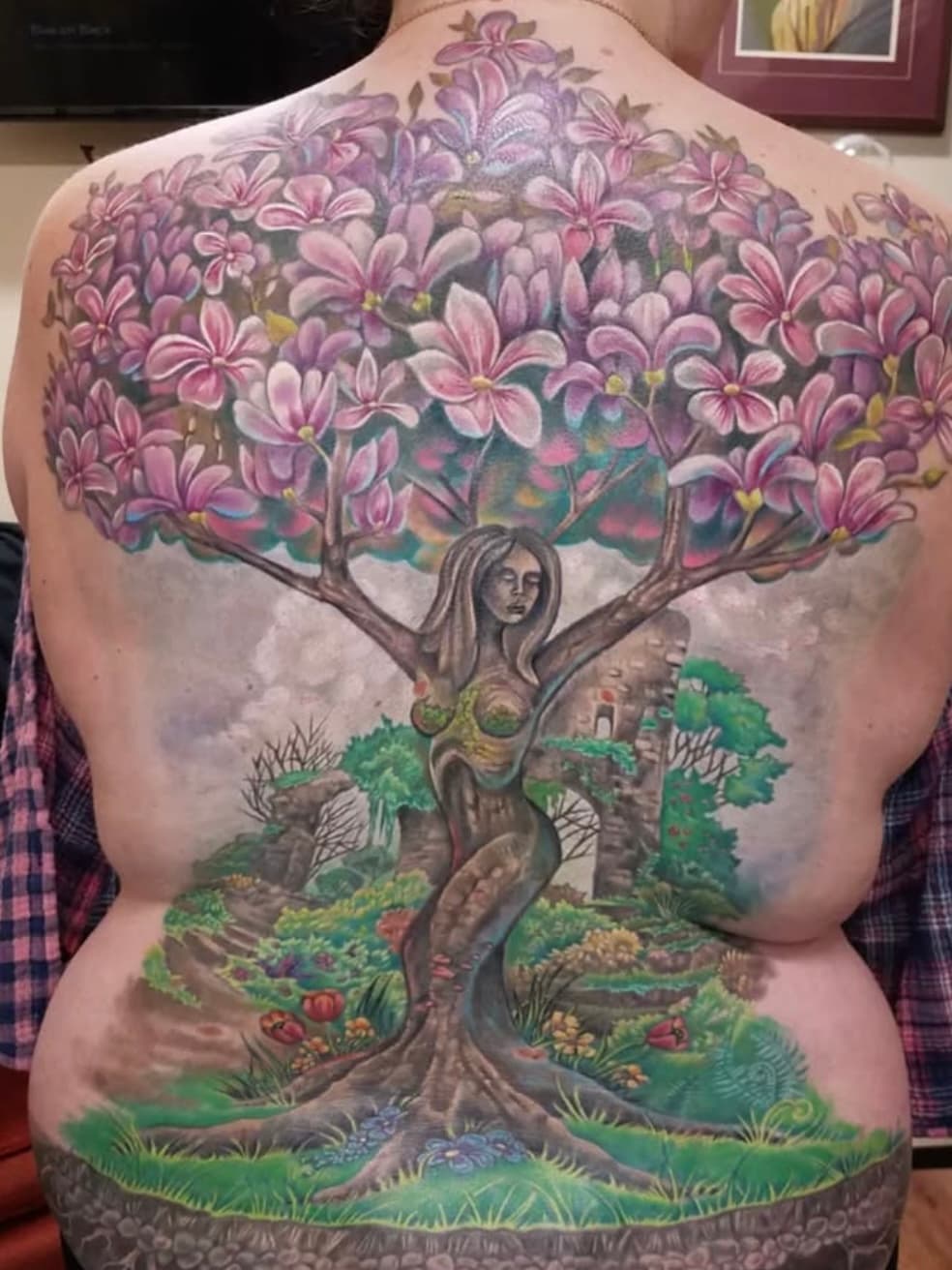 Full back tattoo featuring a woman tree of life with purple blossoms and lush garden landscape
