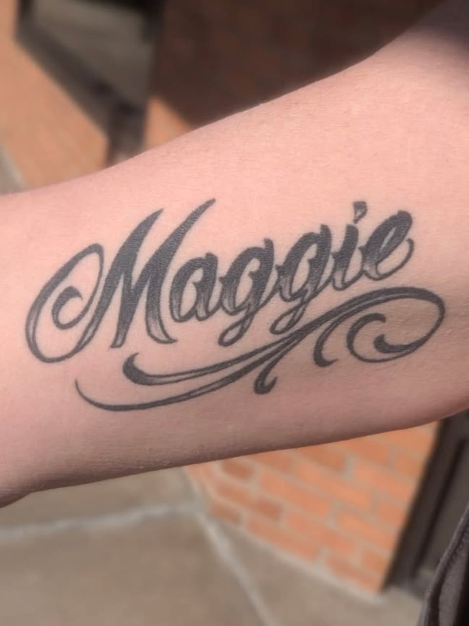 Script tattoo reading 'Maggie' with decorative flourishes on forearm