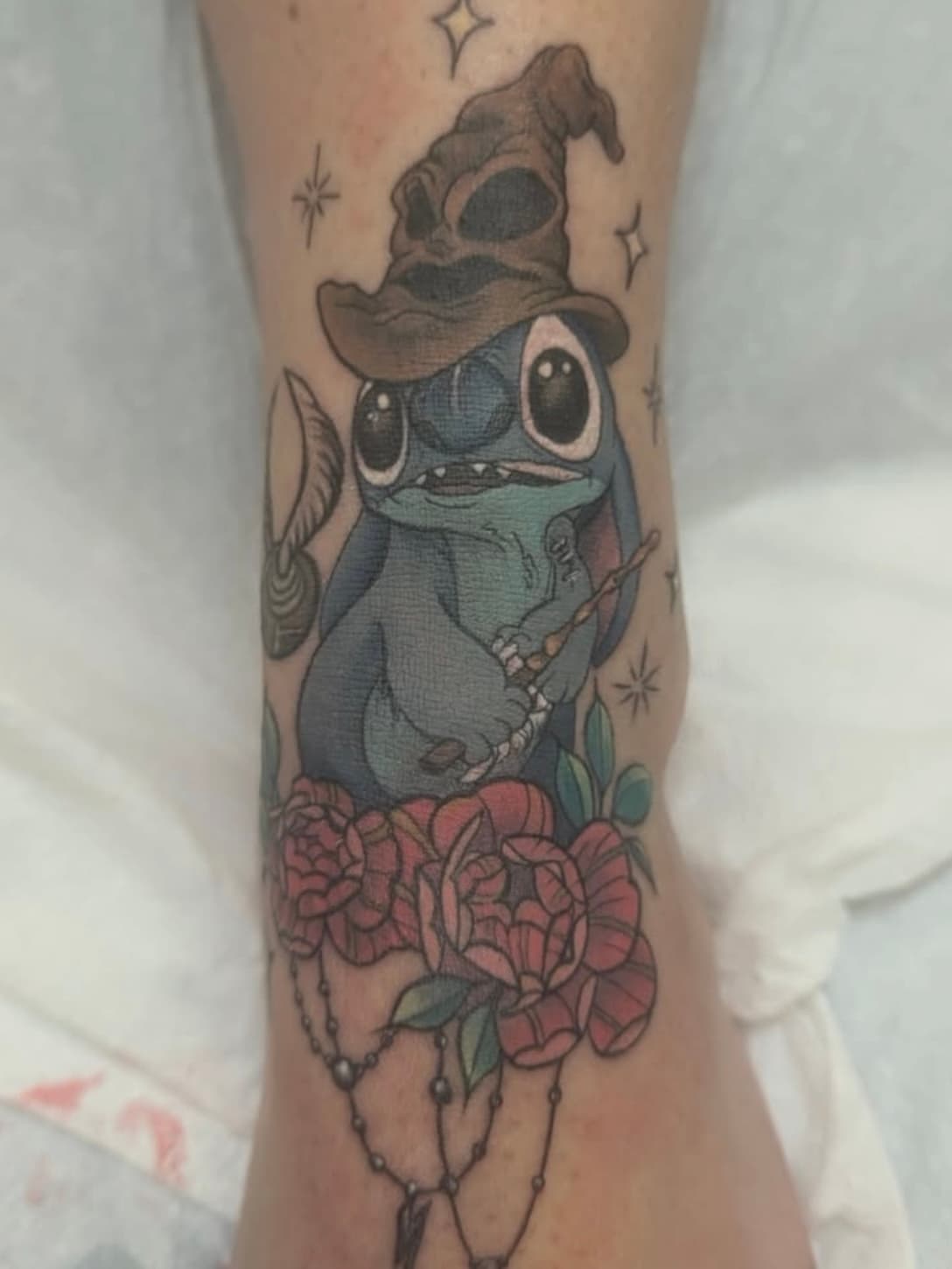 Tattoo of Disney's Stitch character wearing a wizard hat and holding a wand, surrounded by pink roses