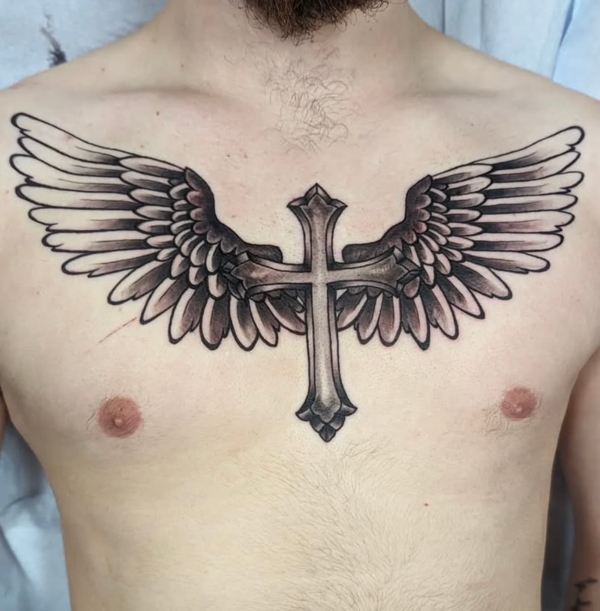 Black and gray tattoo of ornate cross with detailed wings spread across chest